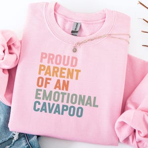 Op de afbeelding: Een lichtroze sweatshirt met de tekst "PROUD PARENT OF AN EMOTIONAL CAVAPOO" in kleurrijke letters. Een delicate gouden ketting met een kleine hanger ligt op de trui. De trui is op een spijkerbroek gevouwen.