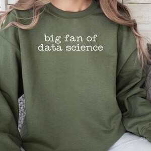 Puede incluir: Una sudadera de cuello redondo en color verde oliva con el texto blanco "big fan of data science" impreso en una fuente estilo máquina de escribir en el pecho. La prenda tiene mangas largas con puños acanalados y un cuello también acanalado.