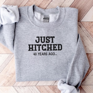 Op de afbeelding: Een grijze sweatshirt met de tekst "JUST HITCHED 40 YEARS AGO..." in zwarte letters. De sweatshirt is opgevouwen op een houten oppervlak.