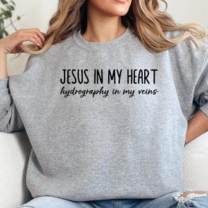 Peut inclure: Un sweat-shirt gris clair avec l'inscription "JESUS IN MY HEART hydrography in my veins" en script noir. Le sweat-shirt à col rond est en matière douce. Le thème général est religieux et inspirant.