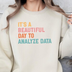May include: Cream-colored sweatshirt with the text "IT'S A BEAUTIFUL DAY TO ANALYZE DATA" in pink, orange, and teal lettering. The sweatshirt has long sleeves and a crew neck.