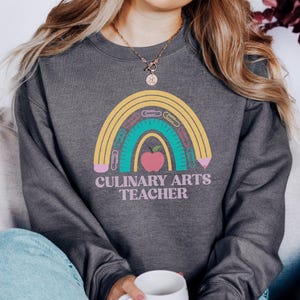 May include: Dark gray sweatshirt with a rainbow design featuring school supplies and a pink apple. The text "CULINARY ARTS TEACHER" is below the rainbow. The sweatshirt is a comfortable, casual style.