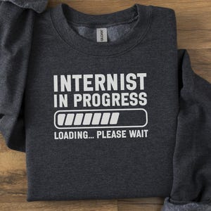 May include: Dark gray sweatshirt with the text "INTERNIST IN PROGRESS" in white, along with a loading bar graphic. Below the graphic, the text reads "LOADING... PLEASE WAIT". The sweatshirt is folded on a wooden surface.
