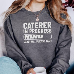 May include: Dark gray sweatshirt with the text "CATERER IN PROGRESS" in white, along with a loading bar graphic and the words "LOADING... PLEASE WAIT." The sweatshirt is a crew neck style.