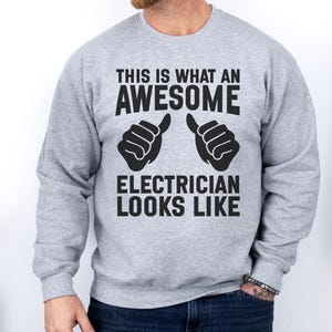 Op de afbeelding: Een lichtgrijs sweatshirt met de zwarte tekst "THIS IS WHAT AN AWESOME ELECTRICIAN LOOKS LIKE". Het ontwerp bevat twee duimen omhoog. De sweatshirt heeft een ronde hals.