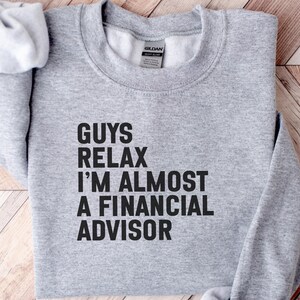 May include: A light grey crewneck sweatshirt featuring the black printed text GUYS RELAX I'M ALMOST A FINANCIAL ADVISOR. The garment has long sleeves, a ribbed collar, and a Gildan brand label visible at the inner neckline of the heathered fabric.