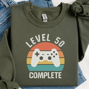 May include: Olive green sweatshirt with a retro design. The graphic features a white game controller, the words "LEVEL 50 COMPLETE", and a sunset-style background. The sweatshirt is folded on top of a pair of blue jeans.