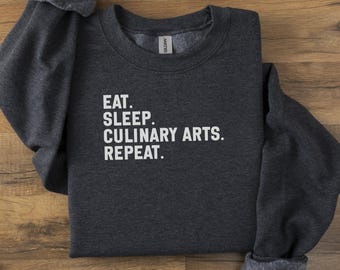 May include: Dark gray sweatshirt with the words "EAT. SLEEP. CULINARY ARTS. REPEAT." in white block letters. The sweatshirt is folded on a wooden surface.