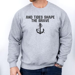 Op de afbeelding: Een lichtgrijs sweatshirt met de tekst "AND TIDES SHAPE THE BRAVE" boven een zwarte ankerafbeelding. Het sweatshirt met lange mouwen heeft een ronde hals. De persoon draagt een blauwe spijkerbroek.