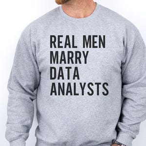 以下が含まれることがあります： 胸元に太字のサンセリフ体で「REAL MEN MARRY DATA ANALYSTS」という黒いテキストがプリントされた、ヘザーグレーのクルーネックトレーナー。長袖で、襟元、袖口、裾はリブ編みになっています。
