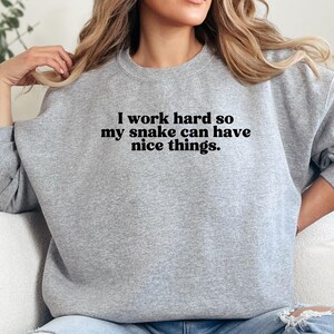Op de afbeelding: Een lichtgrijs sweatshirt met de tekst "I work hard so my snake can have nice things." De tekst is zwart. De trui heeft een ronde hals en lange mouwen.