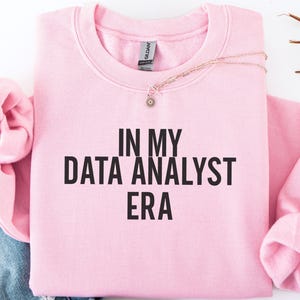May include: A light pink crewneck sweatshirt featuring the black text "IN MY DATA ANALYST ERA" printed in a bold, sans-serif font across the chest. The garment has long sleeves and a ribbed collar, with a small silver-toned necklace resting on top.
