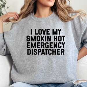 May include: A light gray sweatshirt with the text "I LOVE MY SMOKIN HOT EMERGENCY DISPATCHER" in bold black letters. The sweatshirt has long sleeves and a crew neck.