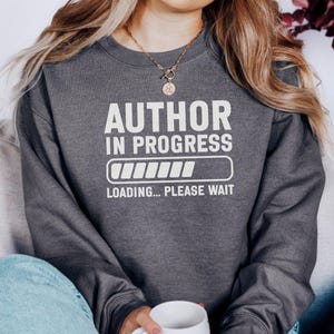 May include: Dark gray sweatshirt with the text "AUTHOR IN PROGRESS" in white, along with a loading bar graphic and the words "LOADING... PLEASE WAIT". The sweatshirt is a crew neck style.