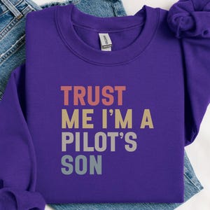 May include: A purple sweatshirt with the text "TRUST ME I'M A PILOT'S SON" in a retro font. The text is in shades of red, tan, and grey. The sweatshirt is folded on top of a pair of blue jeans.