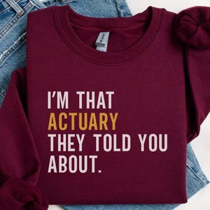 May include: A maroon sweatshirt with the text "I'M THAT ACTUARY THEY TOLD YOU ABOUT." The word "ACTUARY" is in gold. The sweatshirt is folded on top of a pair of blue jeans.