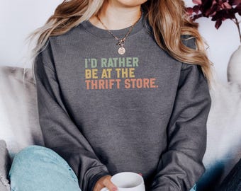 May include: Dark gray sweatshirt with the text "I'D RATHER BE AT THE THRIFT STORE" in green, yellow, and orange. The sweatshirt is a casual style, perfect for everyday wear. The text has a vintage, retro design.
