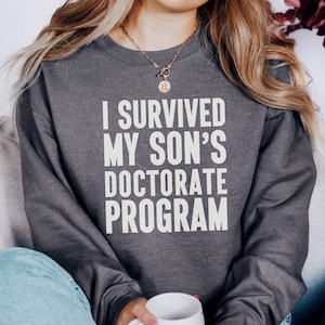 Könnte beinhalten: Dunkelgraues Sweatshirt mit dem weißen Text "I SURVIVED MY SON'S DOCTORATE PROGRAM". Das Sweatshirt hat einen Rundhalsausschnitt. Die Person trägt eine goldene Halskette und hält eine weiße Tasse.
