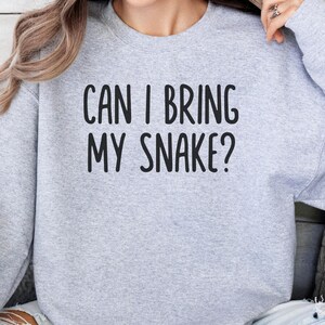 Op de afbeelding: Een lichtgrijs sweatshirt met de zwarte tekst "CAN I BRING MY SNAKE?". De sweatshirt heeft een ronde hals en lijkt gemaakt van een zacht materiaal.