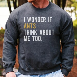 Op de afbeelding: Donkergrijze sweatshirt met de tekst "I WONDER IF ANTS THINK ABOUT ME TOO." Het woord "ANTS" is goudkleurig. De sweatshirt is een crew neck stijl.