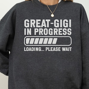 May include: A dark charcoal gray crewneck sweatshirt featuring white text that reads GREAT-GIGI IN PROGRESS above a loading bar graphic and the text LOADING... PLEASE WAIT. The garment has long sleeves and a ribbed neckline, providing a casual look.