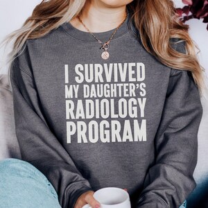 May include: Dark gray sweatshirt with the text "I SURVIVED MY DAUGHTER'S RADIOLOGY PROGRAM" in white block letters. The sweatshirt is worn by a person holding a white mug. The person is wearing blue jeans.