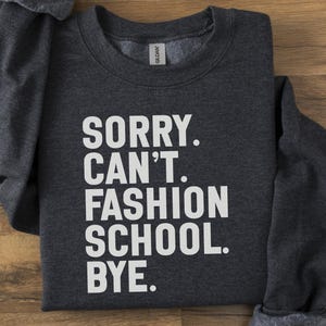 May include: Charcoal grey sweatshirt featuring the white text: "SORRY. CAN'T. FASHION SCHOOL. BYE." The garment is folded on a wooden surface. The Gildan brand label is visible.