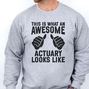 Puede incluir: Sudadera gris claro con el texto negro "THIS IS WHAT AN AWESOME ACTUARY LOOKS LIKE". El diseño incluye dos gestos de manos negras con el pulgar hacia arriba. La sudadera es de cuello redondo.