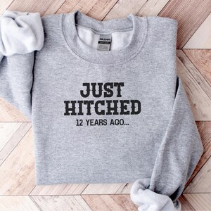Może przedstawiać: Szara bluza z napisem "JUST HITCHED 12 YEARS AGO..." w czarnym kolorze. Bluza jest złożona na drewnianej powierzchni.
