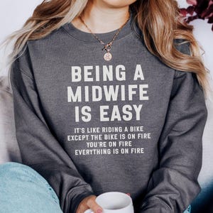 May include: Dark gray sweatshirt with the text "BEING A MIDWIFE IS EASY" in white. Below the main text is the phrase "IT'S LIKE RIDING A BIKE EXCEPT THE BIKE IS ON FIRE YOU'RE ON FIRE EVERYTHING IS ON FIRE".