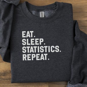 Op de afbeelding: Donkergrijze sweatshirt met de witte tekst "EAT. SLEEP. STATISTICS. REPEAT.". Het kledingstuk is op een houten oppervlak gevouwen. Het Gildan-merklabel is zichtbaar in de kraag.