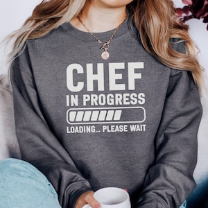 May include: Dark gray sweatshirt with the text "CHEF IN PROGRESS" in white block letters. Below the text is a loading bar graphic. The phrase "LOADING... PLEASE WAIT" is printed below the loading bar.