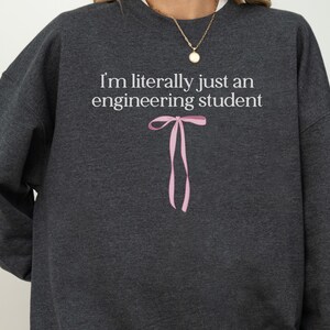 Op de afbeelding: Een donkergrijze sweater met ronde hals en de witte tekst "I'm literally just an engineering student" op de borst, geplaatst boven een eenvoudige, lichtroze grafische illustratie van een gestrikte lintstrik.