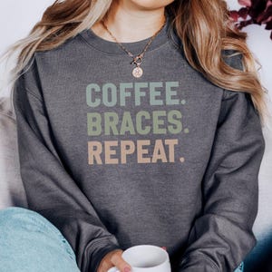 Puede incluir: Sudadera gris oscuro con el texto "COFFEE. BRACES. REPEAT." en verde, azul y melocotón. La sudadera es de cuello redondo. La persona lleva vaqueros azules y sostiene una taza blanca.
