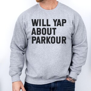 May include: A light gray sweatshirt with the text "WILL YAP ABOUT PARKOUR" in bold, black capital letters. The sweatshirt is a crew neck style and is being worn by a person. The person is wearing dark blue jeans.