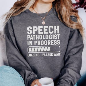 Könnte beinhalten: Dunkelgraues Sweatshirt mit dem weißen Text "SPEECH PATHOLOGIST IN PROGRESS" und einer Ladebalken-Grafik. Der Satz "LOADING... PLEASE WAIT" steht darunter. Das Sweatshirt hat einen lässigen Stil.