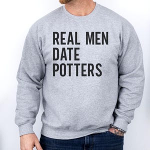 Könnte beinhalten: Hellgraues Sweatshirt mit dem Text "REAL MEN DATE POTTERS" in fetten, schwarzen Buchstaben. Das Sweatshirt hat einen Rundhalsausschnitt. Der Hintergrund ist eine schlichte weiße Wand, rechts hängt eine Jeansjacke.