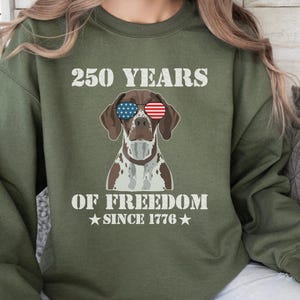 Puede incluir: Sudadera verde oliva con el texto "250 YEARS OF FREEDOM SINCE 1776" y un gráfico de un perro con gafas de sol con diseños de la bandera estadounidense. El perro tiene marcas marrones y blancas.