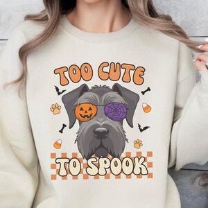 Puede incluir: Una sudadera beige de cuello redondo con el gráfico de un perro gris que lleva gafas con un cristal de calabaza naranja y otro de telaraña morada. El texto TOO CUTE TO SPOOK está rodeado de murciélagos, caramelos y huellas de patas.