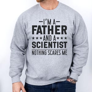 Op de afbeelding: Een lichtgrijs sweatshirt met de tekst "I'M A FATHER AND A SCIENTIST NOTHING SCARES ME" in vette zwarte letters. De sweatshirt heeft lange mouwen en een ronde hals.