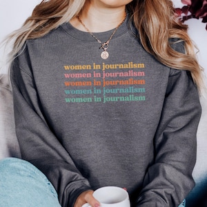 May include: Dark gray sweatshirt with the text "women in journalism" in a stacked design. The text is in a retro color palette of orange, pink, teal, and yellow. The sweatshirt is a casual style.