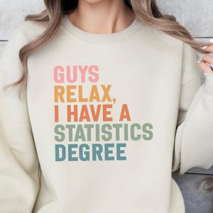 May include: A cream-coloured sweatshirt featuring the text "GUYS RELAX, I HAVE A STATISTICS DEGREE" in a retro font. The text is stacked vertically, with each line in a different colour. The sweatshirt has long sleeves.