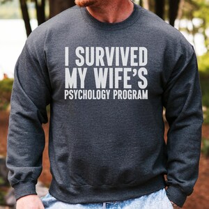 May include: Dark gray sweatshirt with the white text "I SURVIVED MY WIFE'S PSYCHOLOGY PROGRAM." The long-sleeved crew neck shirt is made of a soft material. The background shows trees and a body of water.