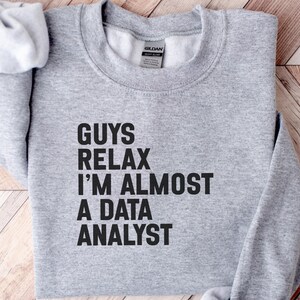 May include: A light gray crewneck sweatshirt featuring the black text "GUYS RELAX I'M ALMOST A DATA ANALYST" printed on the front. The garment has long sleeves, a ribbed collar, and a Gildan brand label visible inside the neckline.