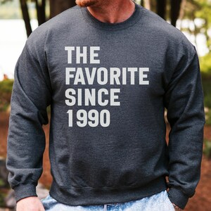 Puede incluir: Sudadera gris oscuro con el texto blanco "THE FAVORITE SINCE 1990". La sudadera es de cuello redondo y parece estar hecha de un material suave. La imagen está tomada al aire libre.