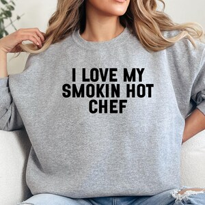 May include: A light gray sweatshirt with the text "I LOVE MY SMOKIN HOT CHEF" in bold black letters. The long-sleeved crew neck sweatshirt is a casual style.