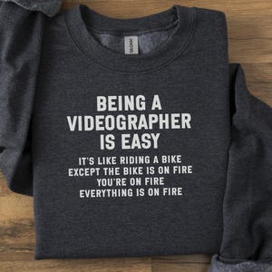 May include: Charcoal grey sweatshirt featuring the white text: "BEING A VIDEOGRAPHER IS EASY". The humorous quote below reads: "IT'S LIKE RIDING A BIKE EXCEPT THE BIKE IS ON FIRE YOU'RE ON FIRE EVERYTHING IS ON FIRE."