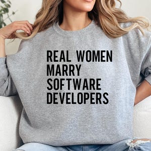 May include: A pale grey sweatshirt featuring the black text "REAL WOMEN MARRY SOFTWARE DEVELOPERS". The sweatshirt has a crew neck and long sleeves.