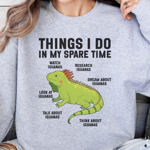 May include: A light gray crewneck sweatshirt featuring a central graphic of a green iguana. The text reads: THINGS I DO IN MY SPARE TIME, with labels pointing to the iguana: WATCH IGUANAS, RESEARCH IGUANAS, DREAM ABOUT IGUANAS, LOOK AT IGUANAS, TALK ABOUT IGUANAS, THINK ABOUT IGUANAS.