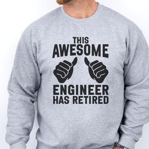May include: A light gray sweatshirt with the text "THIS AWESOME ENGINEER HAS RETIRED" in bold black letters. The design includes two black thumbs-up symbols. The sweatshirt is a crew neck style.